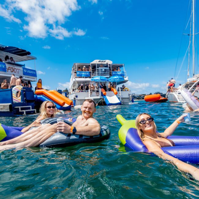 People relax on inflatable loungers and floaties in the water near several anchored boats at a lively outdoor party under a bright blue sky. Many are smiling and enjoying drinks in the sun.