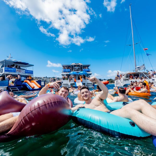 Two men smile and cheers drinks while floating on inflatable loungers in the water, surrounded by other people enjoying a lively boat party under a bright blue sky.