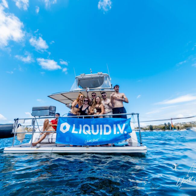 A group of people in swimwear stand and pose on the deck of a boat with a blue "LIQUID I.V." banner, surrounded by water under a partly cloudy sky.