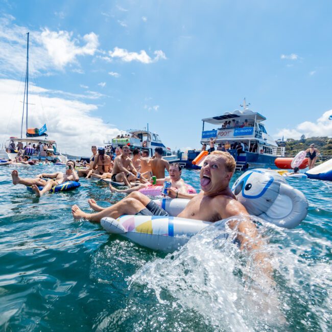 A group of people enjoy a lively party on the water, relaxing on colorful inflatable floats near boats. One man in the foreground laughs and splashes water, while others socialize under a sunny blue sky.