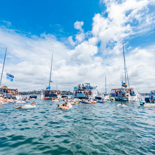 People swim and float on inflatables in a busy, festive lake surrounded by boats under a partly cloudy sky. The scene suggests a lively summer gathering or party on the water.