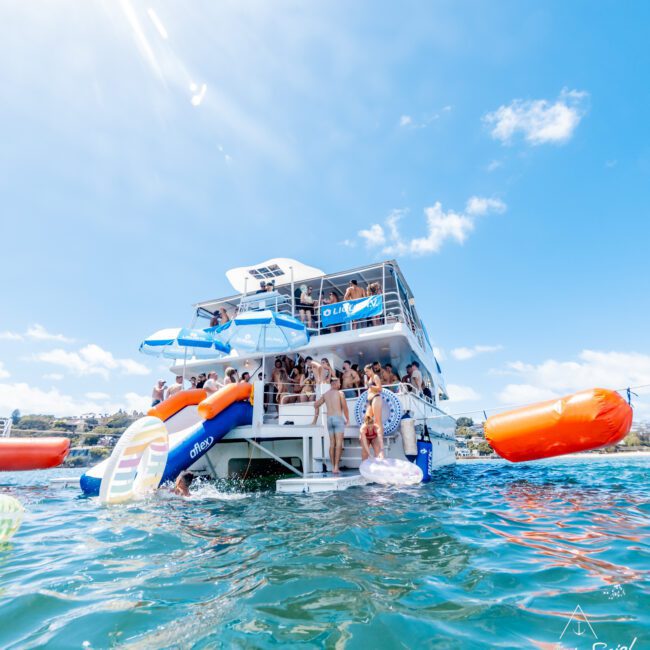 A lively group of people enjoy a sunny day on a double-decker boat with slides into the water, large floaties, and blue skies above. The boat is surrounded by clear, inviting water.