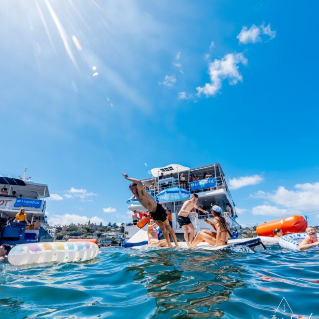 People enjoy a sunny day on the water, with some diving off a floating platform, boats in the background, and others relaxing on inflatables. The image is vibrant, with blue skies and sun rays overhead.