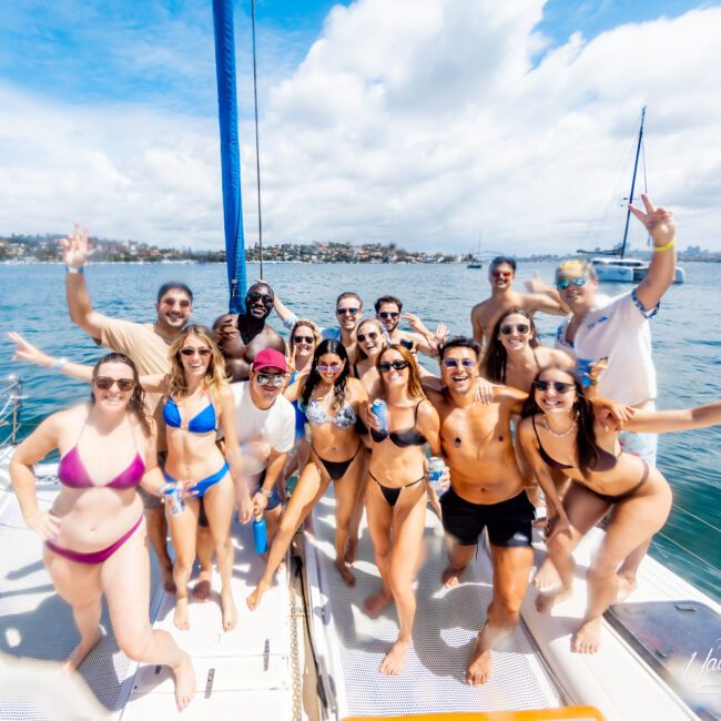 A group of people in swimsuits smile and pose together on a boat under a sunny sky, with blue water and another sailboat in the background.