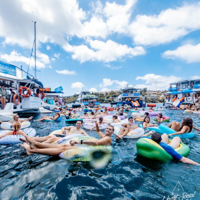 A lively group of people relax on colorful inflatables in the water, surrounded by boats and yachts under a partly cloudy sky. The scene is festive and social, with many enjoying the sunshine and the water.