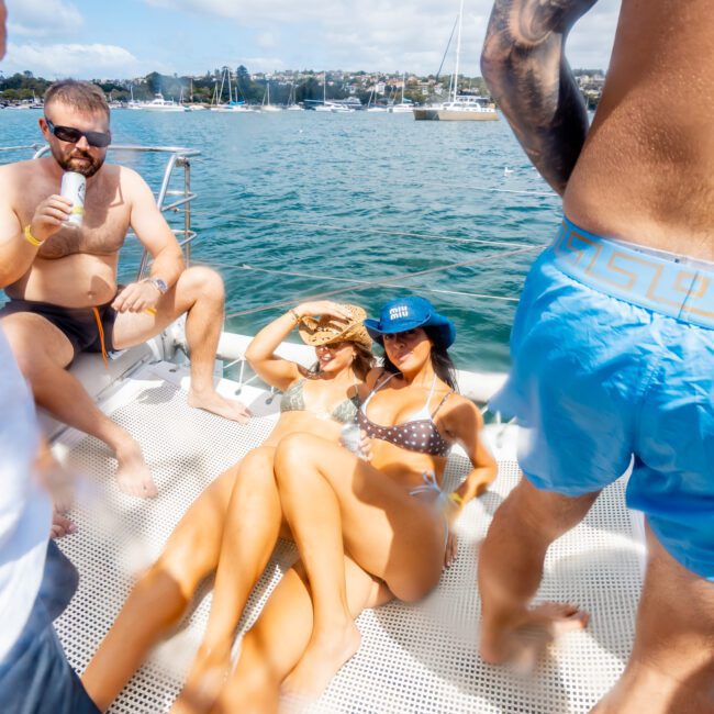 Two women in bikinis relax and pose on a boat deck, wearing sunglasses and hats, while others in swimwear stand and sit nearby. The boat is on the water with sailboats and a cityscape in the background.