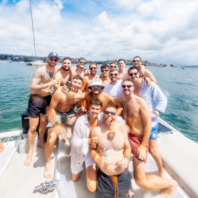 A group of smiling men in swimwear pose together on the deck of a boat, holding drinks, with blue sky, clouds, and water in the background. It looks like a fun, sunny day on the water.