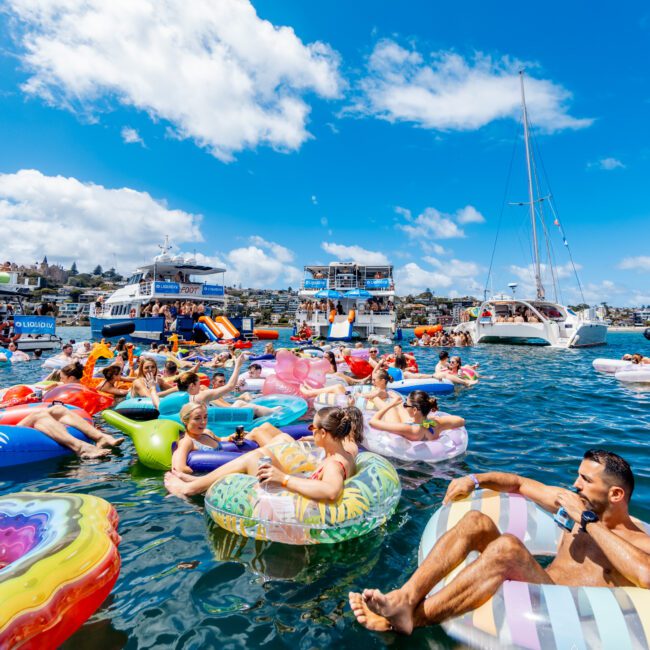 A group of people relaxing on colorful inflatable floats in a busy, sunlit harbor with boats and yachts in the background under a bright blue sky with scattered clouds.