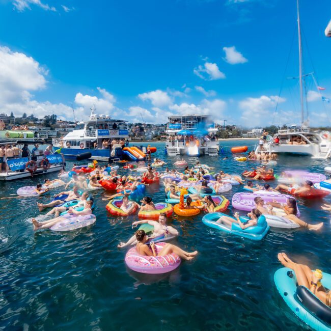 A lively scene of people relaxing on colorful inflatable tubes in the water near several anchored boats under a bright blue sky, with a coastal town visible in the background.