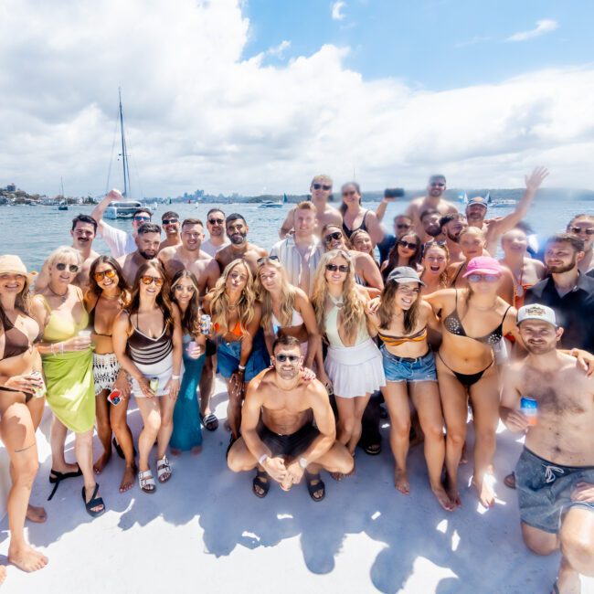 A large group of people in swimsuits and summer attire pose and smile on the deck of a boat, with water, yachts, and a distant shoreline in the background under a partly cloudy sky.