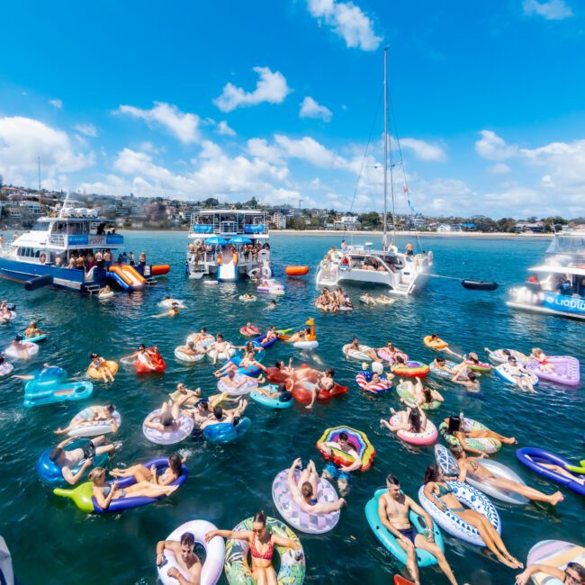 People relaxing on colorful inflatable floats in a sunny blue harbor, surrounded by several boats and yachts. The sky is bright with scattered clouds, creating a vibrant and festive summer atmosphere.