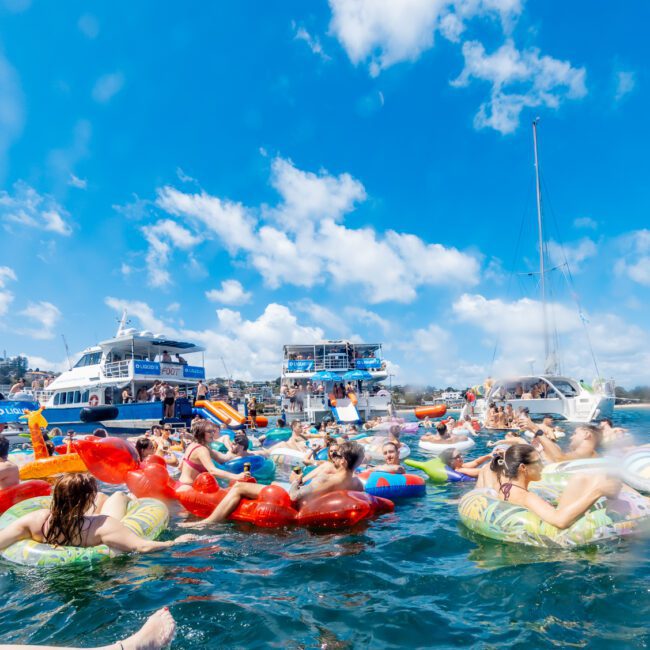 A large group of people relax on colorful inflatables in the water, surrounded by boats under a sunny blue sky with scattered clouds. A bird flies overhead, and the scene is lively and festive.
