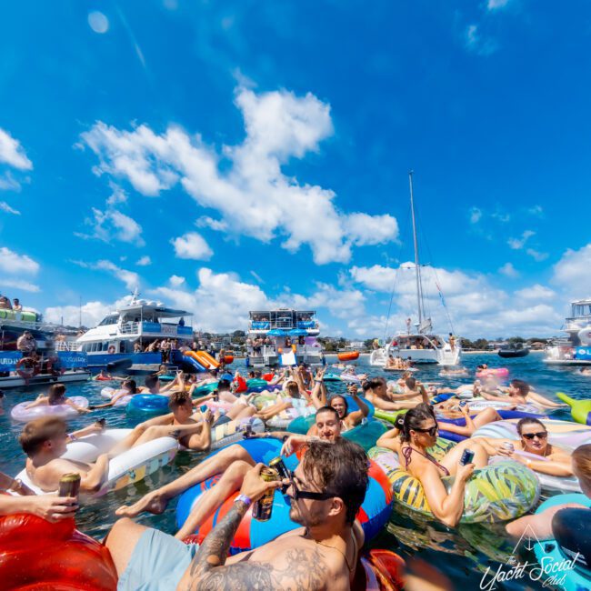 A large group of people relaxing on colorful inflatables in the water, holding drinks, with boats and a bright blue sky in the background at a lively outdoor event.