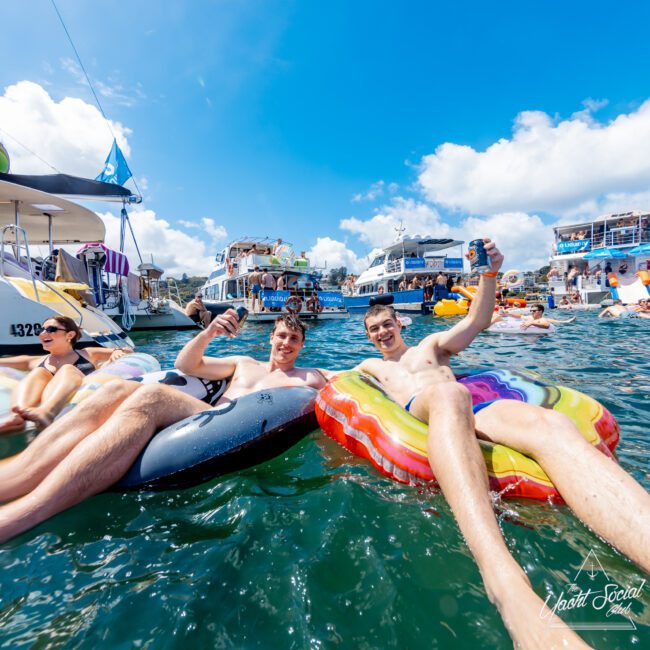 Two young men relax on colorful inflatable floats in the water, smiling and holding drinks, with boats and other people partying in the background under a bright blue sky.