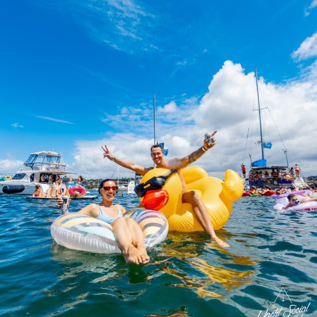 Two people enjoy relaxing on inflatable floats in the water, surrounded by other people and boats. The sky is bright with a few clouds, and the atmosphere is festive and lively.