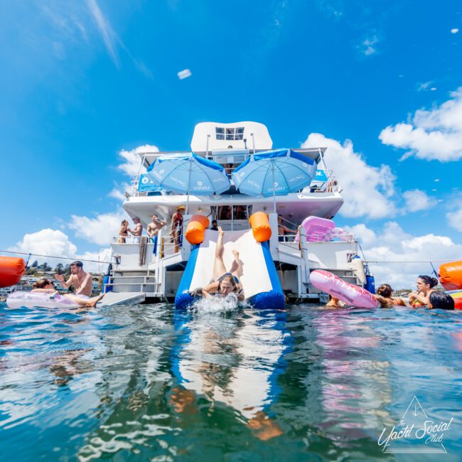 People enjoy a sunny day on a yacht, some swimming in the water and others relaxing on deck. A woman slides down an inflatable slide into the sea, surrounded by floating pool toys and friends. The sky is clear and blue.