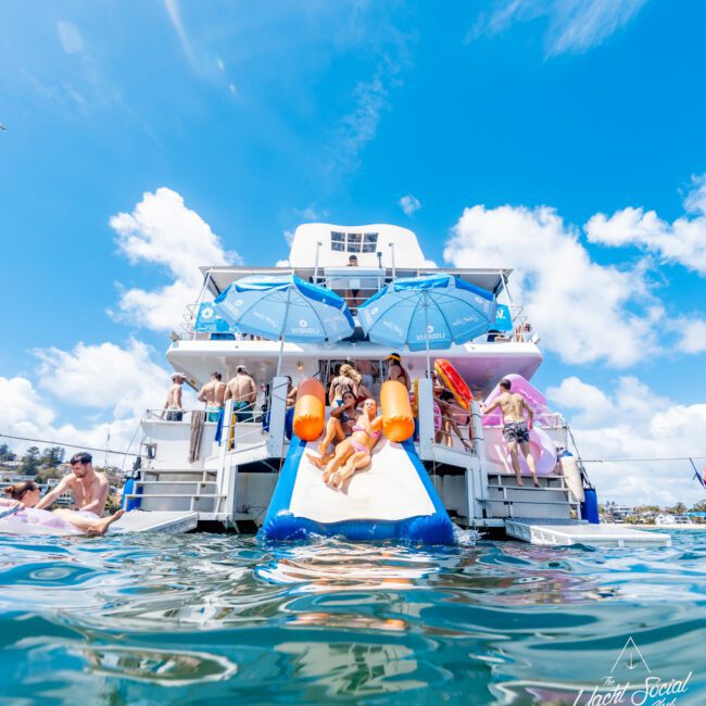 A lively yacht party scene with people in swimwear enjoying sun and water slides on the back of a yacht, under blue umbrellas, with bright skies and water in the foreground. "Yacht Social Club" logo is visible at the bottom right.