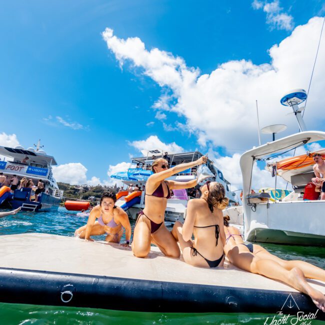 A group of young women in swimsuits relax and play on a floating platform between boats in a sunny, tropical setting. Other people and yachts are visible in the background under a bright blue sky.