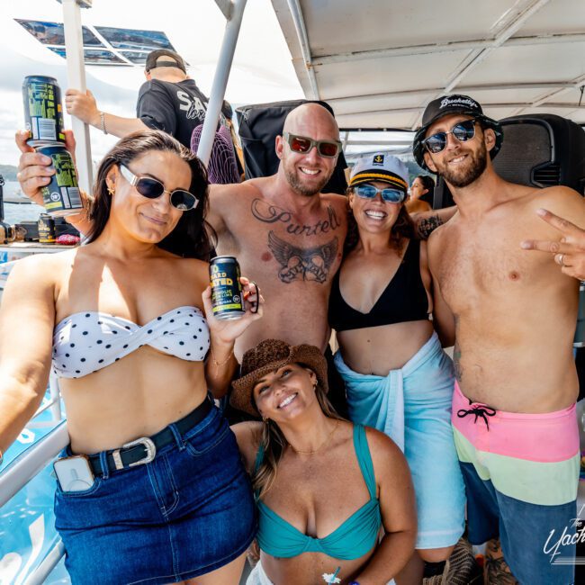 A group of five smiling adults in swimsuits pose together on a boat at a lively outdoor party, holding drinks. Sunlight, water, and other partygoers are visible in the background.