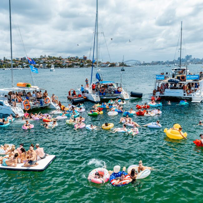 A lively scene of people partying on boats and colorful inflatables in a harbor, with city buildings and the Sydney Harbour Bridge visible in the background under a cloudy sky.