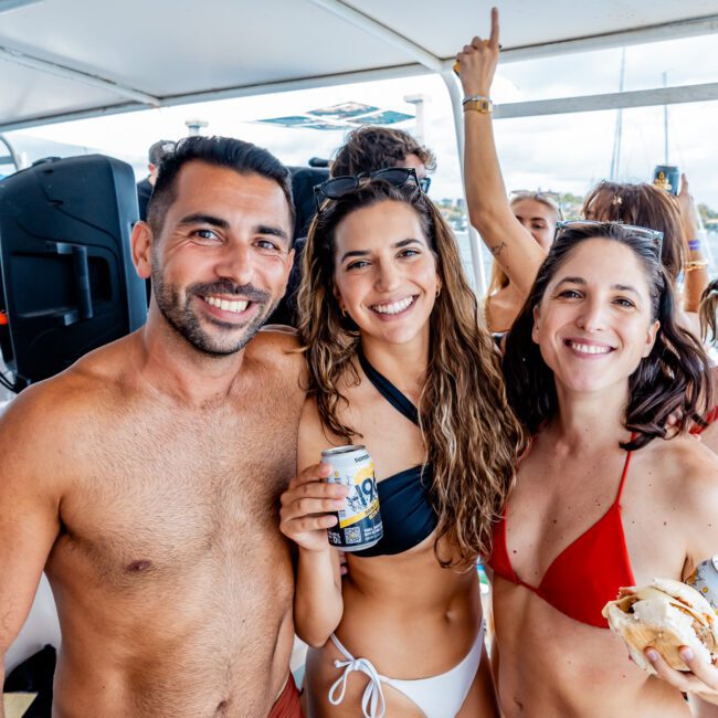 Three people in swimwear smile at the camera on a boat; two women hold drinks and a sandwich, and the man stands to the left. Other people and boats are visible in the background, suggesting a lively party atmosphere.