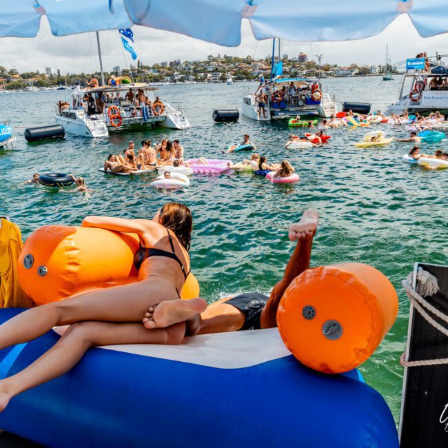Two people relax on an inflatable raft at a lively boat party, surrounded by other people on floats and boats in the water under a sunny sky.