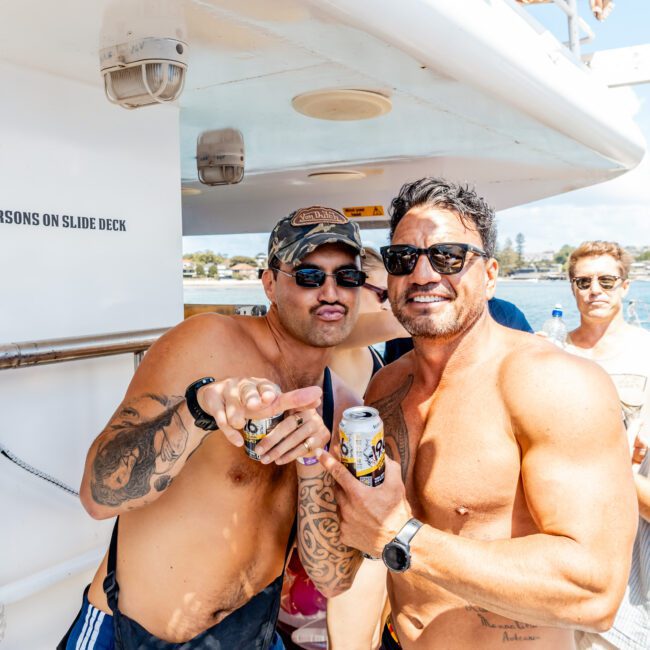 Two shirtless men with sunglasses smile and pose with drinks on a sunny boat deck. Other people and parts of the boat are visible in the background.