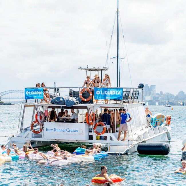 A large party boat with people dancing and relaxing is anchored in a bay, surrounded by people floating on colorful inflatables in the water. A city skyline and bridge are visible in the background under a cloudy sky.