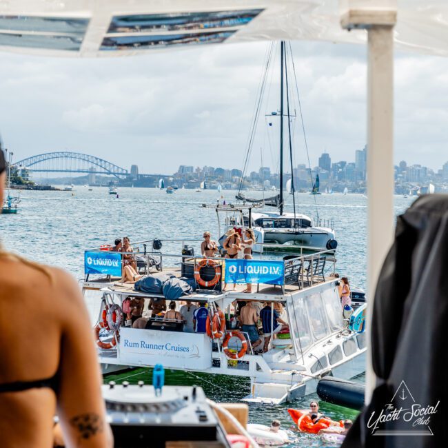 A group of people party on docked yachts in a harbor, with banners reading "LIQUID I.V." and "Rum Runner Cruises." The Sydney Harbour Bridge and city skyline are visible in the background.