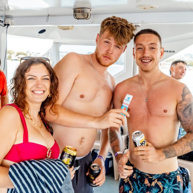 Three friends in swimwear smile and pose on a boat, holding drinks and sunscreen. The woman holds a striped towel, while the two men stand beside her, one with his arm around the other. Other people and boat details are visible in the background.