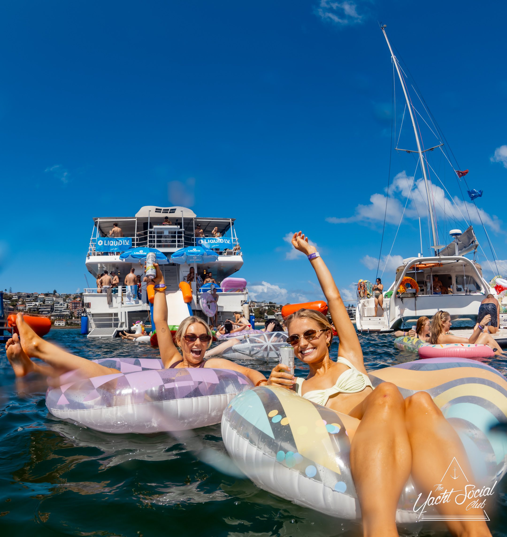 Two women in swimsuits relax on colorful pool floats in the water, smiling and raising their arms. Behind them, people socialize on boats under a clear blue sky. Logo "Yacht Social Club" is visible in the corner.