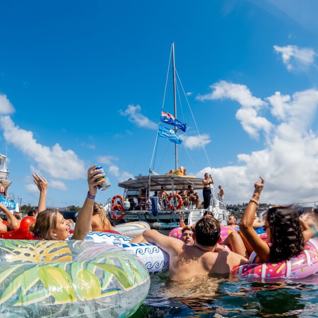 A group of people relax on colorful pool floats in the ocean, raising drinks and enjoying a sunny day near anchored boats with others mingling and music playing in the background. The sky is bright with scattered clouds.