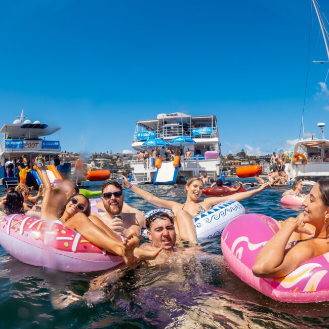 A group of people float on colorful inflatables in the water, smiling and celebrating near yachts and boats on a sunny day, with blue sky and city buildings in the background.