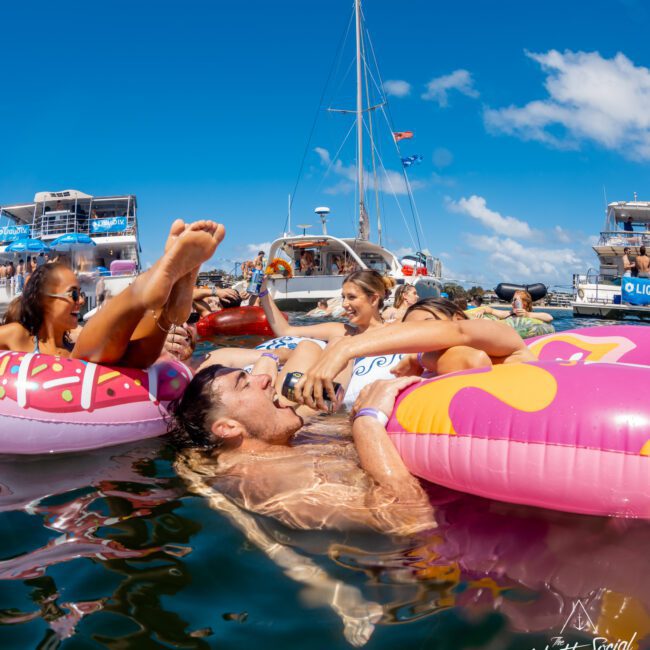 A group of people relax on colorful inflatable floats in the water near yachts under a bright blue sky. They are laughing and enjoying drinks, creating a lively, festive atmosphere.