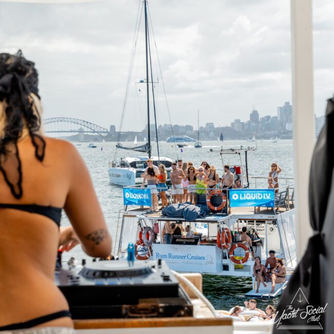 A woman in a swimsuit is DJing on a boat, while a group of people party on another boat nearby. The harbor and city skyline are visible in the background under a cloudy sky.