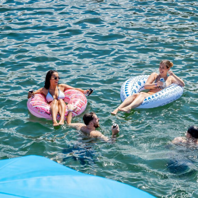 Two women relax on inflatable pool floats, holding drinks, while two men swim nearby in clear blue water. The scene is bright and sunny, with a blue inflatable partially visible in the foreground.