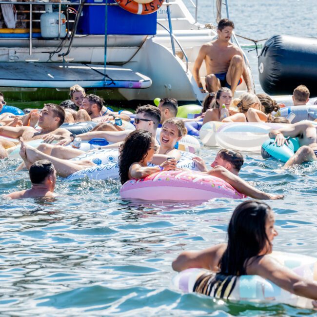 A group of people relax on colorful floaties in the water near a docked boat, with the Sydney Harbour Bridge visible in the background on a sunny day.