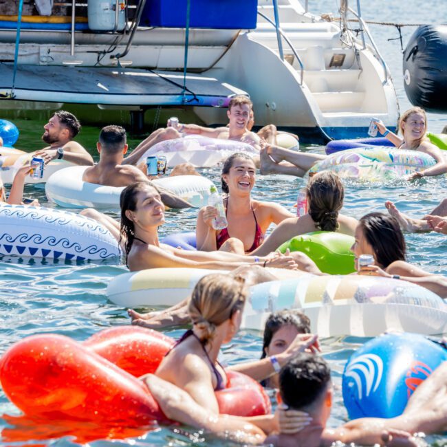 People relax and have fun on colorful inflatable floats in the water near a docked boat on a sunny day, holding drinks and enjoying the lively summer atmosphere.