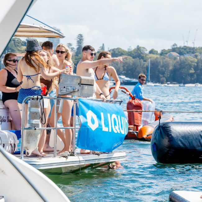 A group of people in swimsuits socializing on a boat decorated with a blue "LIQUI" banner, with some people in the water and the background showing trees and other boats on a sunny day.