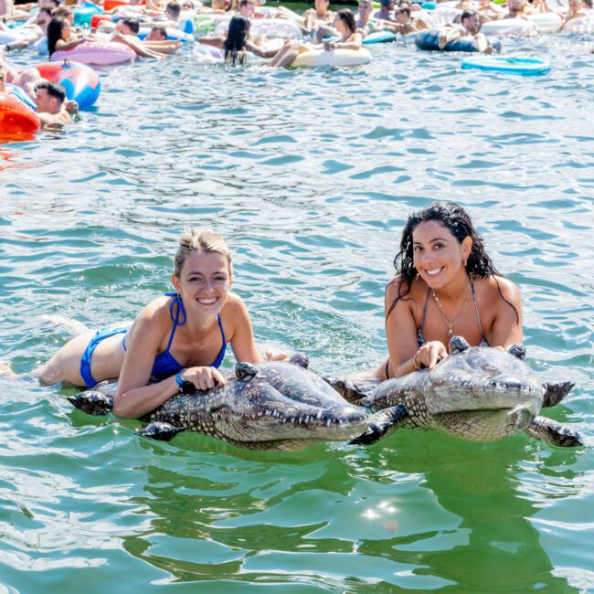 Two women in swimsuits smile at the camera while holding inflatable crocodiles in the water, surrounded by other people on floats, with boats and a bridge visible in the background.