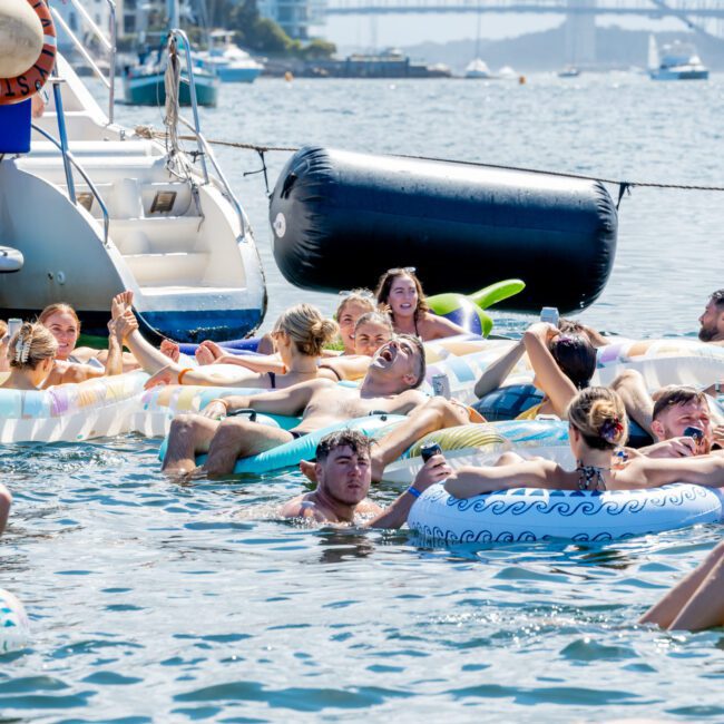 People relaxing on colorful inflatables in the water near a docked boat, enjoying a sunny day. A large bridge and cityscape are visible in the background. The scene is lively and festive.
