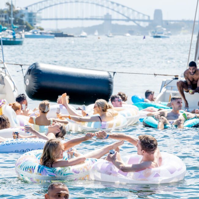 People relax on colorful inflatable tubes in the water near boats, with the Sydney Harbour Bridge visible in the background on a sunny day.
