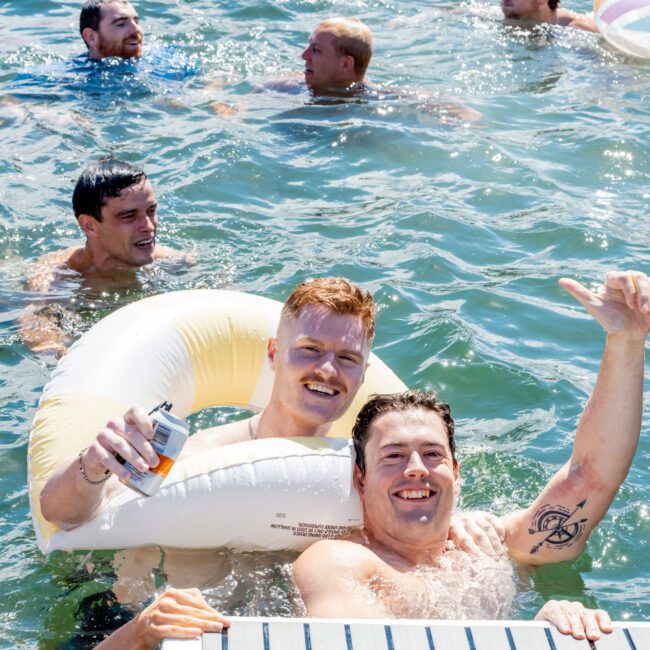 Two men smile for the camera while floating in a lake, one with an inflatable ring and holding a drink. Several others swim and relax on floaties in the sunny water around them. A dock is visible in the foreground.