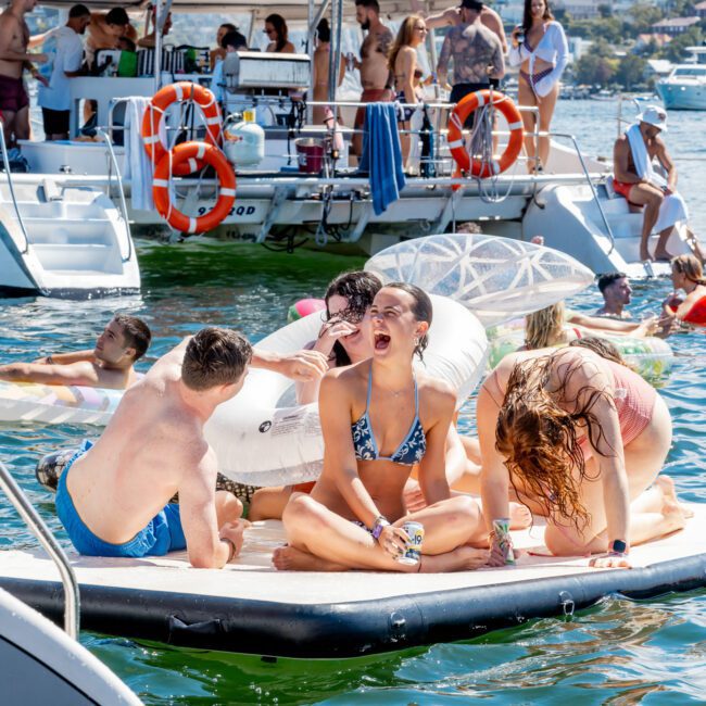 A group of people in swimsuits relax and laugh on a floating mat near boats during a sunny day on the water, with others socializing and swimming in the background.