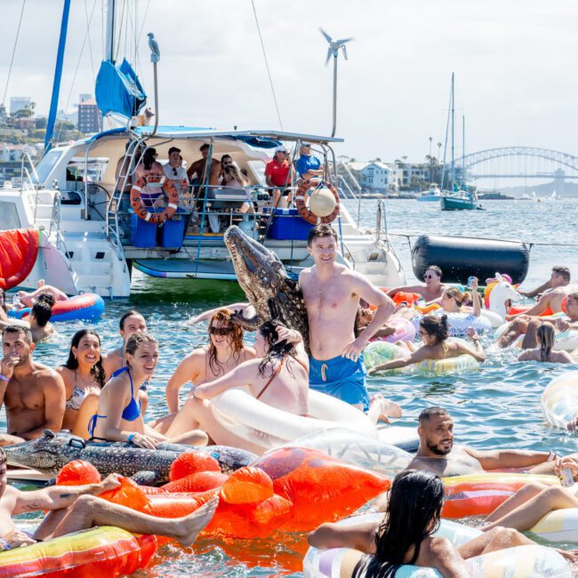 A crowd of people on colorful inflatable floats enjoy a party in the water near boats, with one person holding a large inflatable alligator. The scene is lively and festive under a sunny sky.