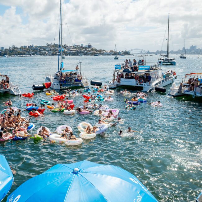 A lively scene of people floating on colorful inflatables and swimming in a bay, surrounded by anchored boats filled with partygoers. Blue umbrellas are in the foreground, and a city skyline is visible in the background.