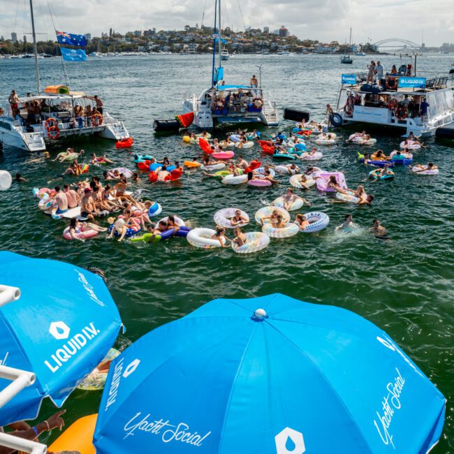 A lively group of people relax on colorful inflatable floats in the water between boats, with large blue "Liquid I.V." umbrellas in the foreground and a cityscape visible across the bay under a partly cloudy sky.