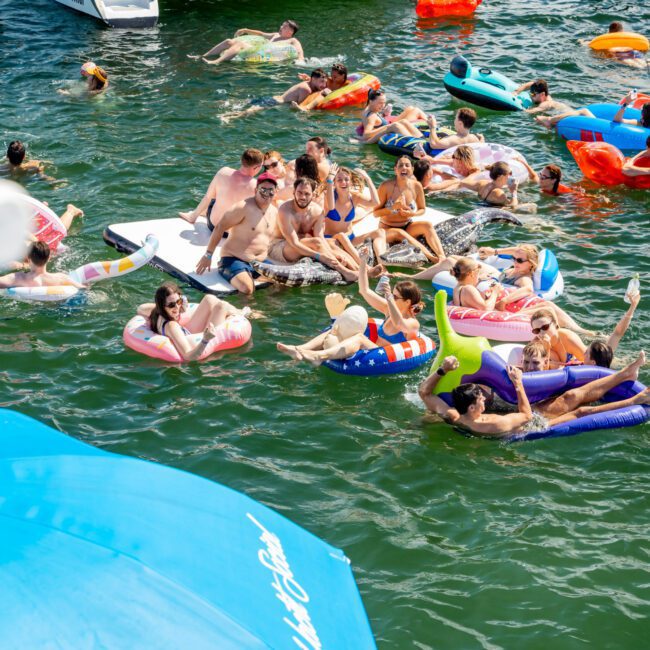 A group of people float on colorful inflatables and lounge near boats in a lively lake party scene, enjoying the sun and water. A large blue umbrella and the logo "Yacht Social Club" are visible in the foreground.