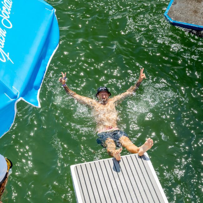 A man in swim trunks joyfully slides off a dock into green water, raising both hands in peace signs. An umbrella and floating platform are visible nearby under bright sunlight.