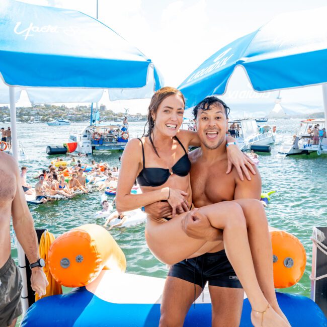 A man in swim trunks playfully lifts a smiling woman in a black bikini as they pose together at a lively pool party on the water, with boats and people swimming in the background under blue umbrellas.